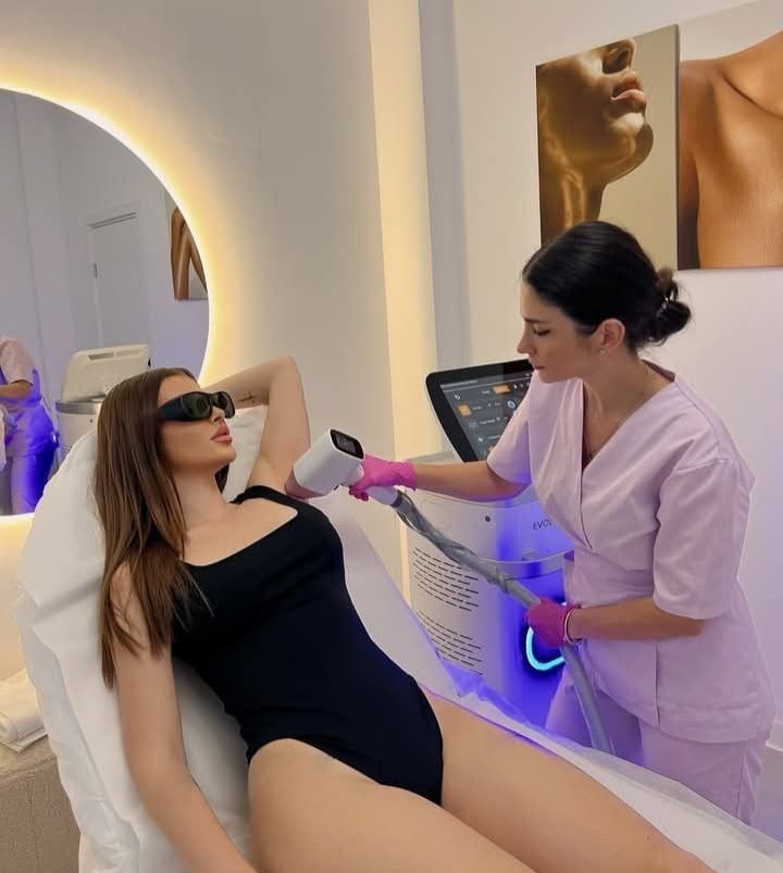 Woman receiving laser hair removal treatment on underarm from aesthetician in modern clinic with blue light device