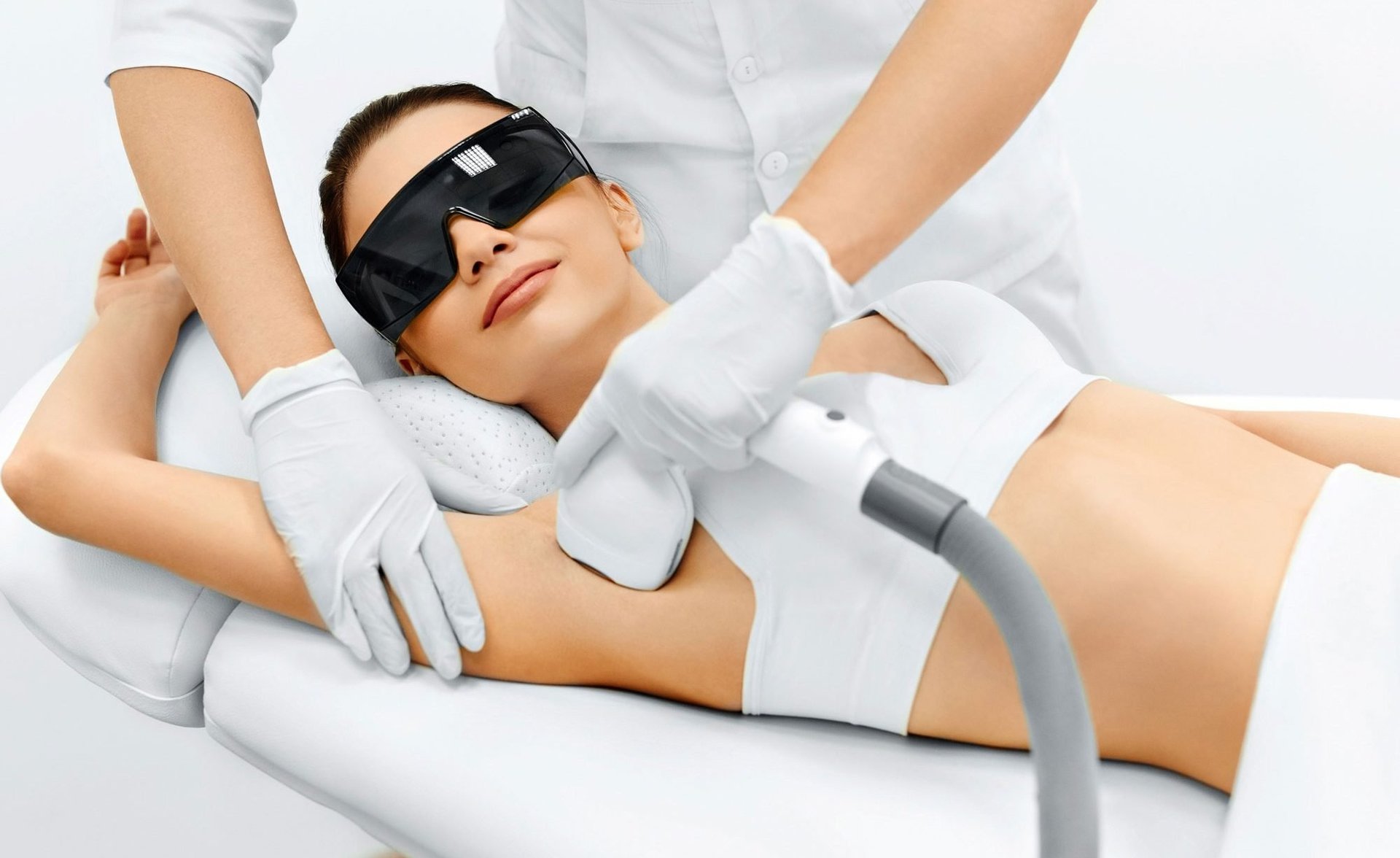 Woman receiving laser hair removal treatment on arm, wearing protective eye goggles and white towel